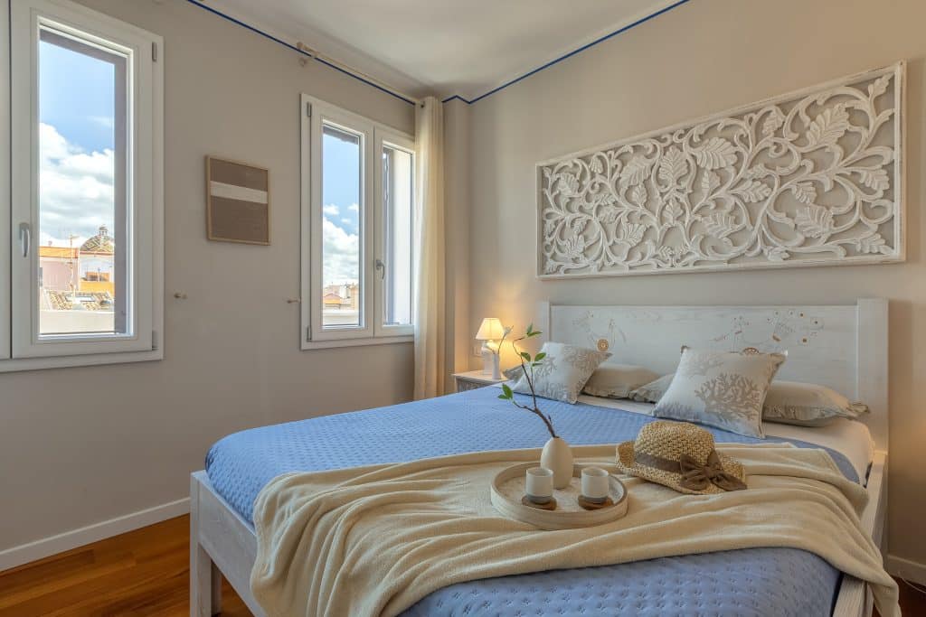Cozy bedroom of an Alghero apartment in Sardinia with handcrafted headboard depicting birds of fertility and a view of San Michele dome. Camera da letto accogliente di un appartamento ad Alghero con testata intagliata a mano raffigurante uccelli della fertilità e vista sulla cupola di San Michele, Sardegna.