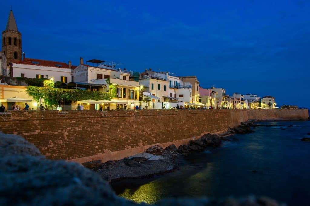 Evening lights along the Alghero old town promenade, filled with restaurants, bars, and walking tourists, with the ForRest Seaside Loft among the historic seafront buildings. Luci serali lungo la passeggiata del centro storico di Alghero, tra ristoranti, bar e turisti che passeggiano, con il ForRest Seaside Loft tra gli edifici storici sul mare.