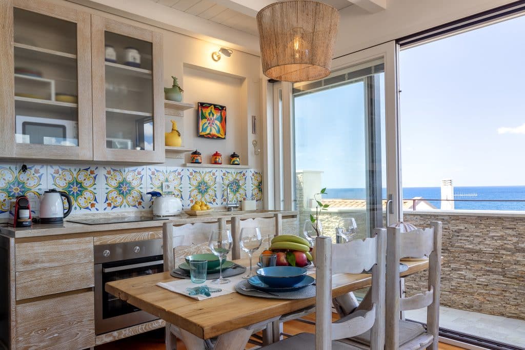 Fully equipped sea-view kitchen and dining area with Sardinian Ginepro wood table, hand-painted tiles and Mediterranean light at ForRest Seaside Loft Alghero Cucina completamente attrezzata con vista mare e zona pranzo con tavolo in legno di ginepro, piastrelle sarde dipinte a mano e luce mediterranea al ForRest Seaside Loft di Alghero